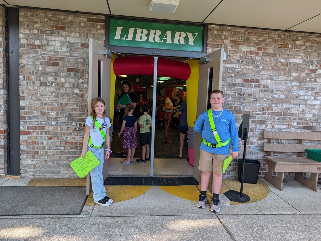 Two children, one in green and the other in blue, stand outside a library with an inflatable entrance. A brick wall, benches, and people are visible.