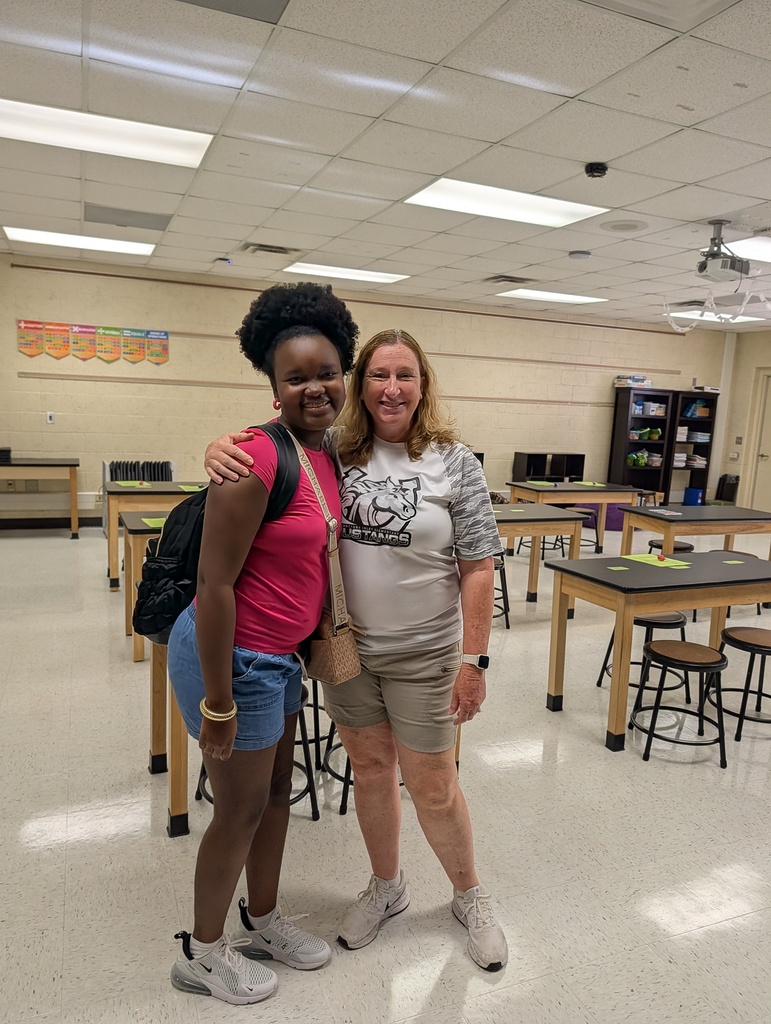 Two women pose for a picture inside a classroom, surrounded by tables and chairs. One wears a pink shirt, shorts, and sneakers.