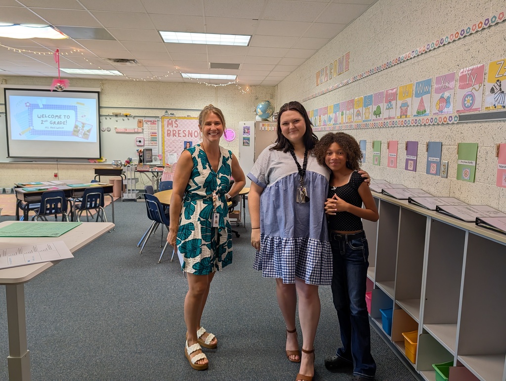 Three women in a classroom, one holding a projector screen. Colorful charts on walls and desks.