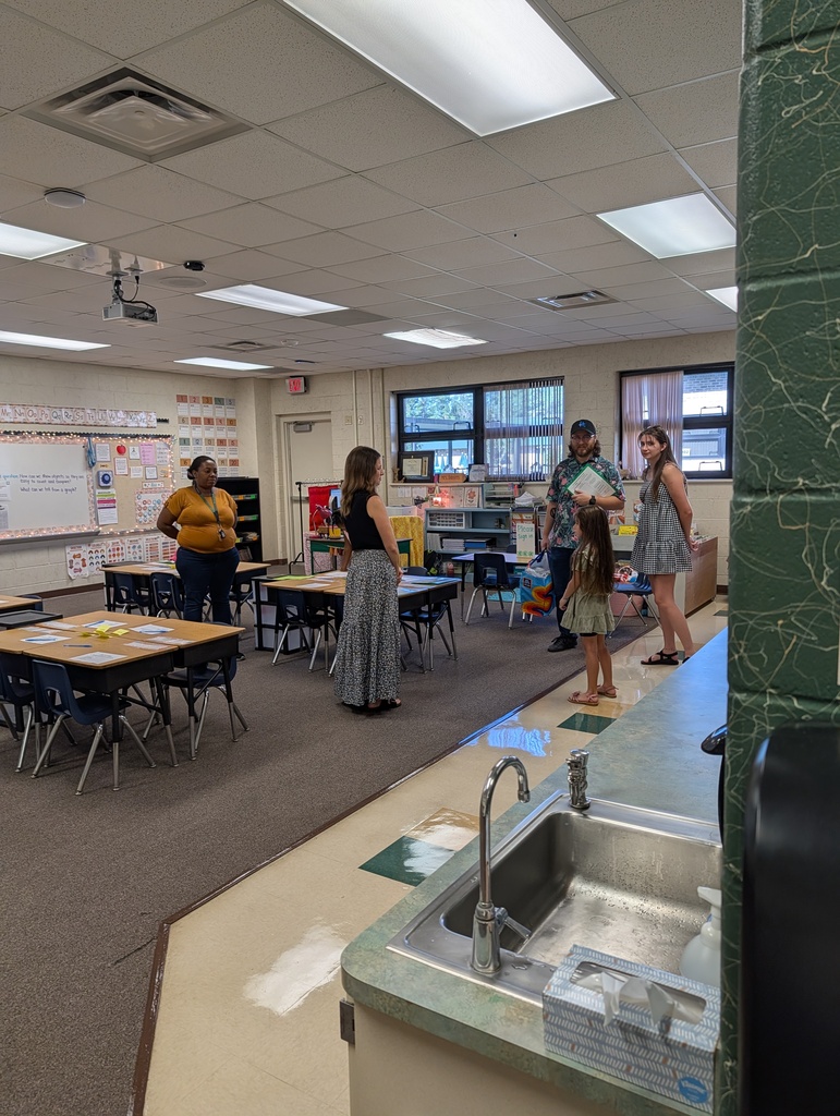 A classroom with desks, chairs, and people standing. Two adults and two children near the tables. A sink with a faucet on the right.