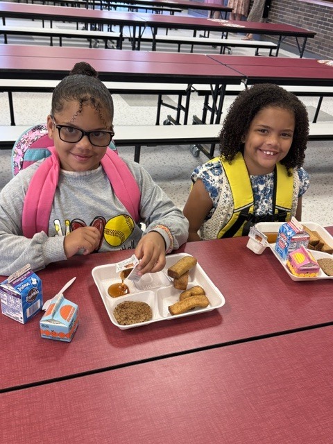 Two children in a cafeteria, seated with trays of food. One has glasses and a backpack, the other wears a yellow vest.