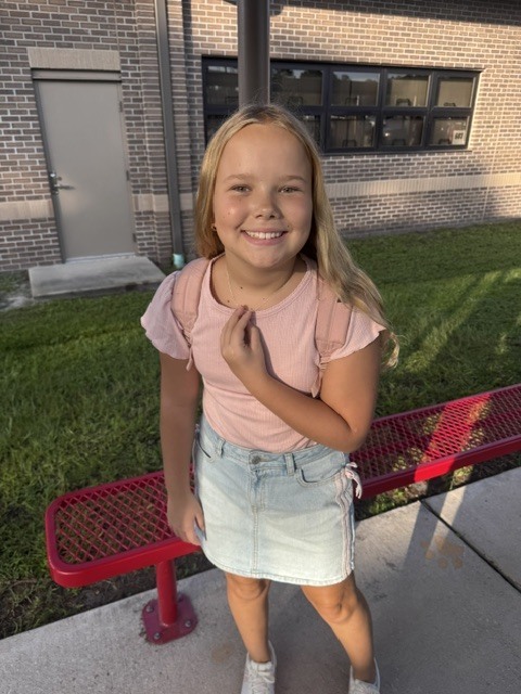 A young girl with blonde hair smiles in front of a red bench, near a building with brick walls.