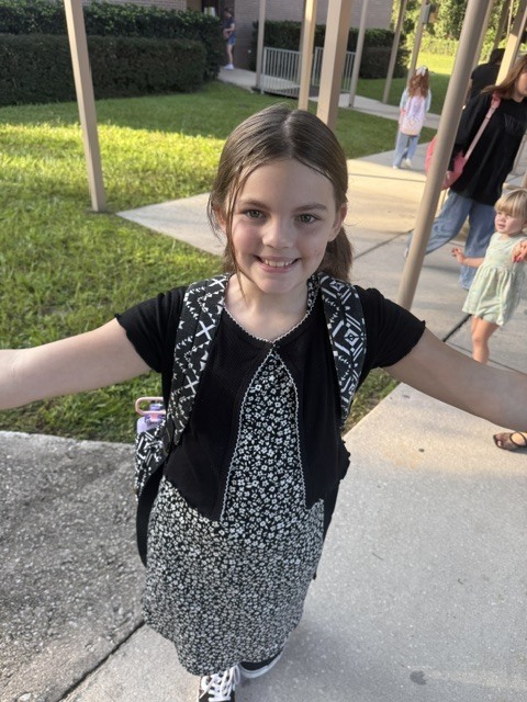 A girl with arms outstretched, wearing a black and white dress and backpack, stands on a sidewalk.