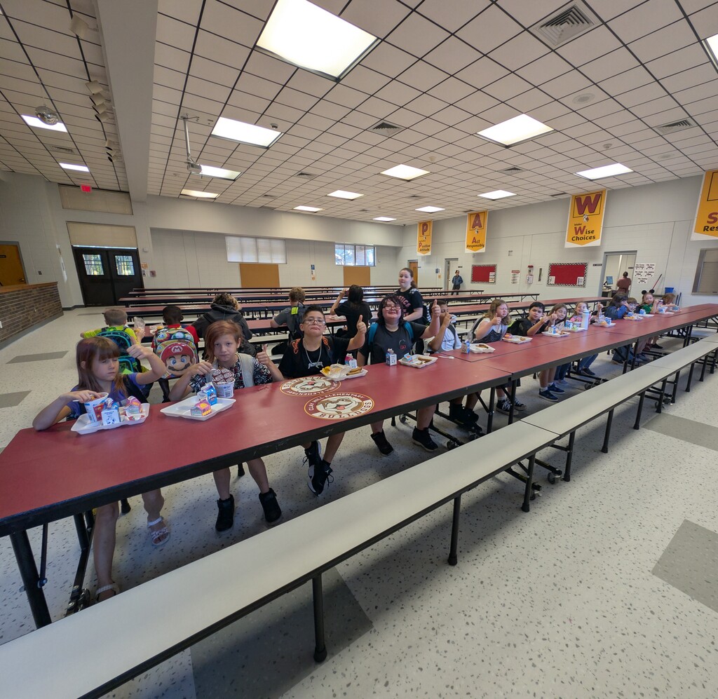 Children seated at tables in a school cafeteria. Red tables and white benches. Bright lights on ceiling.