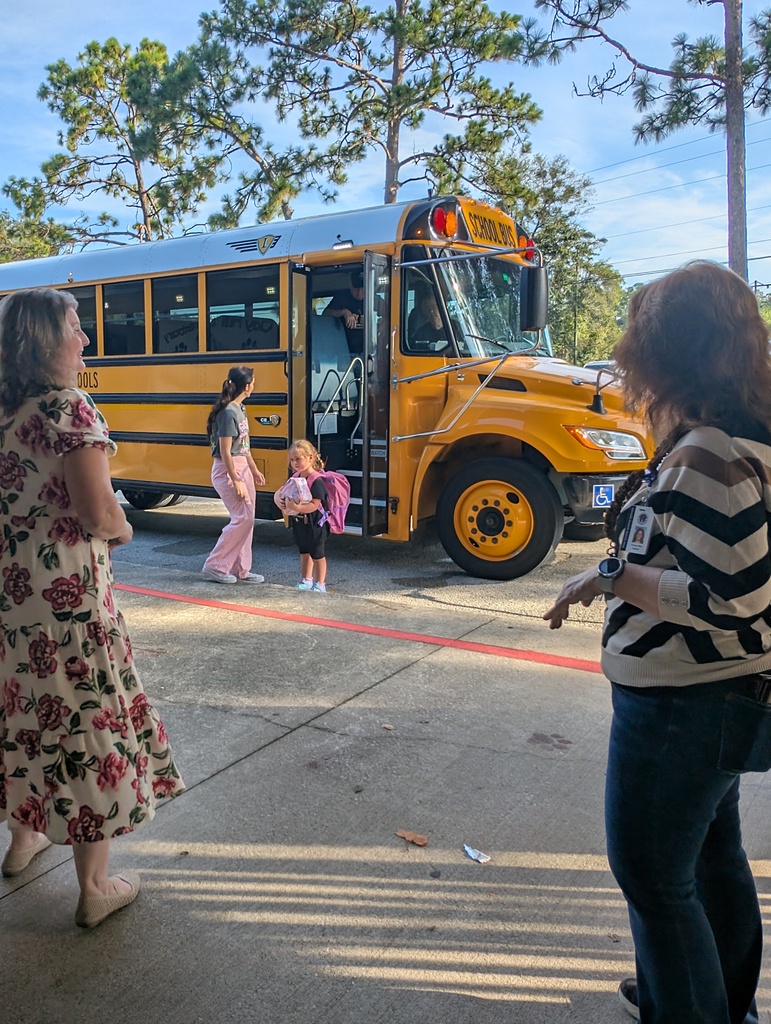 Several people wait by a school bus in a parking lot with trees in the background.