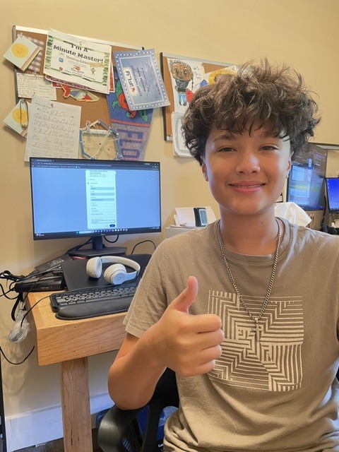 A boy with curly hair gives a thumbs up in a room with a computer desk.