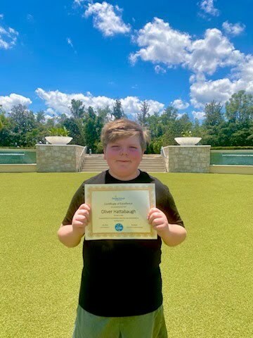 A child holds a certificate in front of a green lawn, with trees and a cloudy sky in the background.