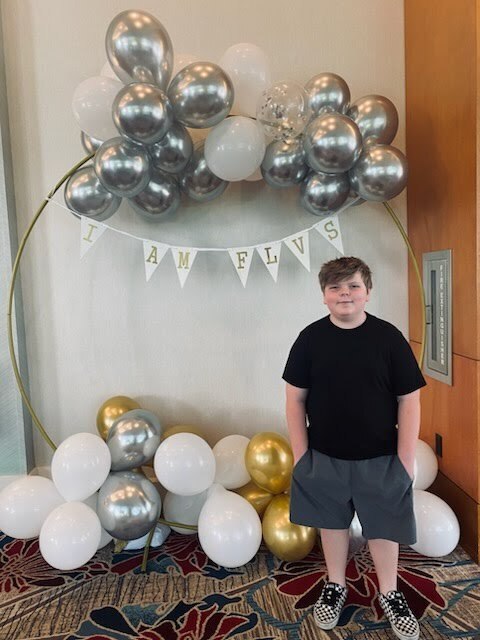 A boy stands in front of a balloon arch and banner with text "I AM FLY".