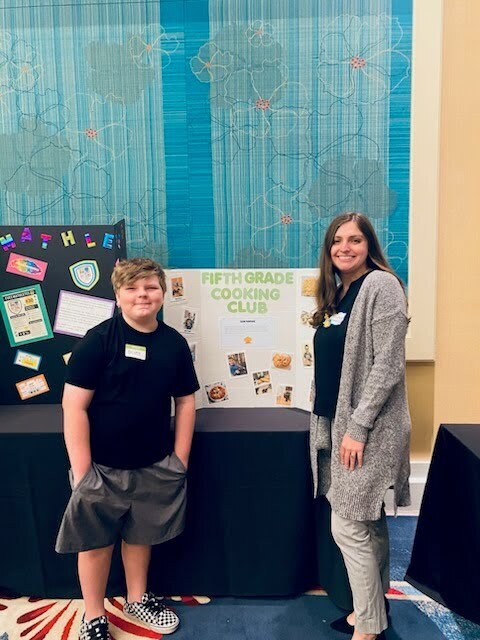 Boy and woman stand before a table. The table displays a bulletin board with the title "Fifth Grade Cooking Club."
