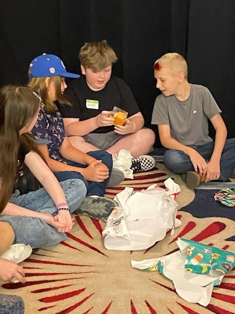 Four young individuals sit on the floor, surrounded by unwrapped presents. One holds a gift, while another has red dye on their head.