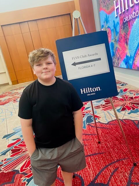 A young boy stands in front of a Hilton sign, wearing a black shirt and shorts. Behind him is a colorful wall and a blue sign pointing to Florida.