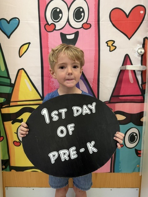 Young child holding a sign in front of a colorful classroom wall with crayons and hearts.