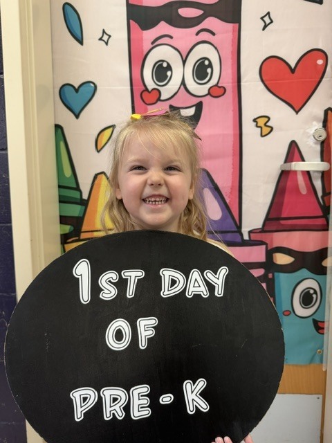A young girl stands in front of a door holding a blackboard that reads "1st Day of Pre-K."