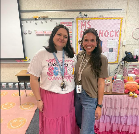 Two women stand in a classroom. One wears a white shirt and pink skirt, the other a brown shirt and jeans.