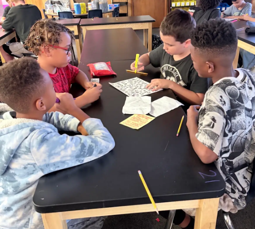 Four boys sit around a table in a classroom, with papers and pencils. One boy uses a red marker.