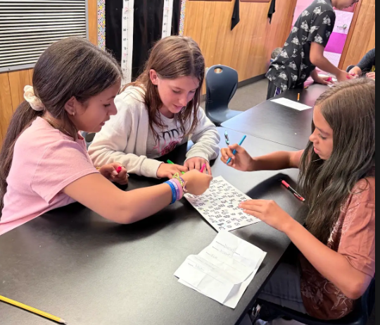 Three children at a table, each holding a pen and working on a worksheet. Two others are nearby.