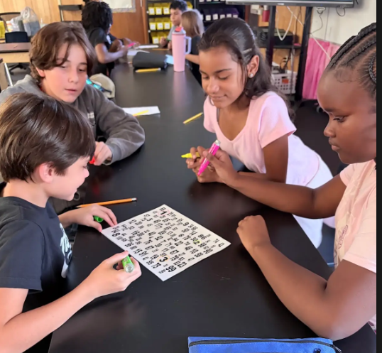Three boys seated at a table, engaging in a puzzle. One boy wears glasses and a black skull shirt.
