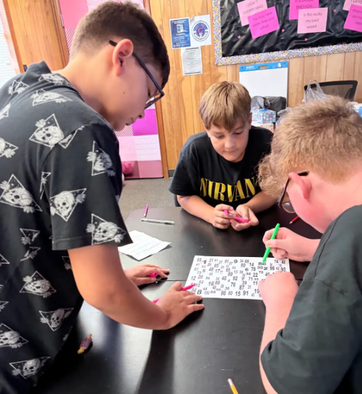 Three boys seated at a table, engaging in a puzzle. One boy wears glasses and a black skull shirt.