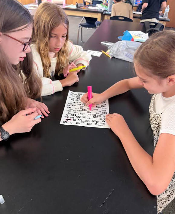 Three young girls seated at a table, using pink markers on a worksheet. Background shows a classroom.
