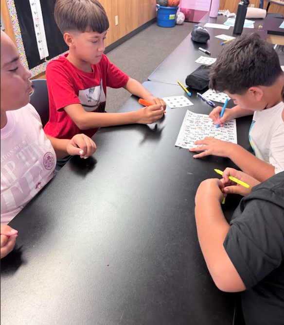 Three boys seated at a table, engaging in a puzzle. One boy wears glasses and a black skull shirt.