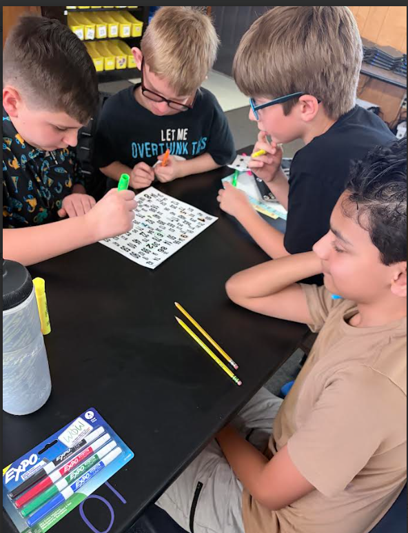 Three boys seated at a table, engaging in a puzzle. One boy wears glasses and a black skull shirt.