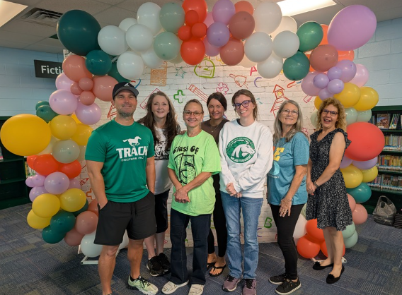People stand behind a colorful balloon arch in a library. They wear various shirts and footwear.