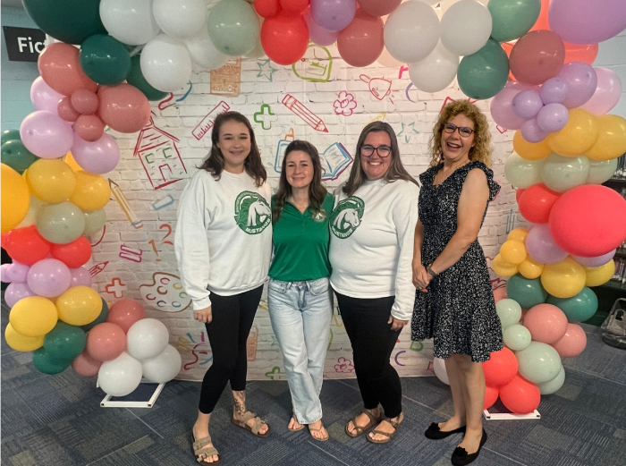 Four women in light-colored tops stand under a colorful balloon arch against a background of educational drawings.