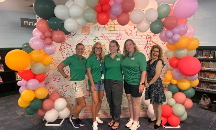 Five women in green shirts stand behind a colorful balloon arch in a library.