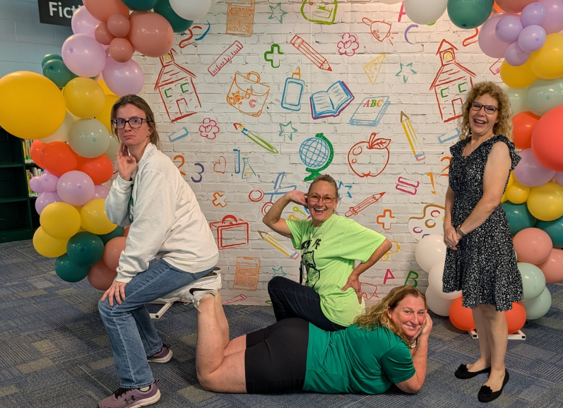 Four women are posing in front of a wall with school-themed drawings and balloons.