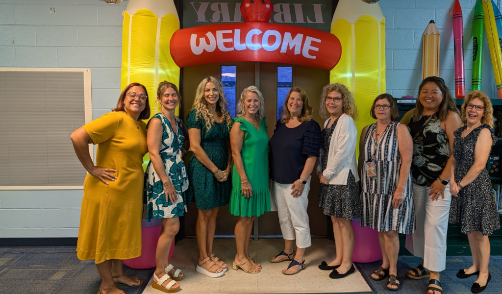 Five women in green shirts stand behind a colorful balloon arch in a library.
