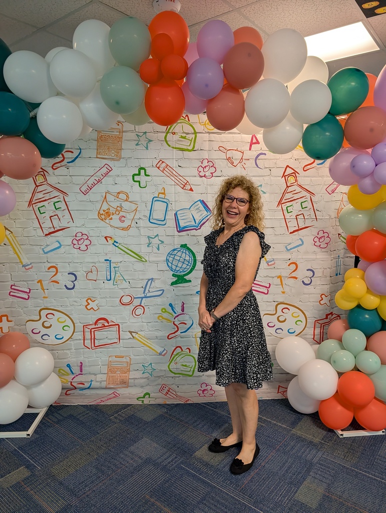 Woman in black dress stands before wall with balloons and images of books, pencils, a globe.
