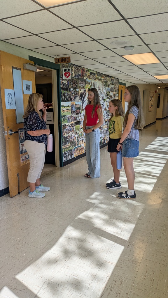 Four individuals stand in a hallway, two wearing glasses, one wearing sandals. One woman holds a pink object. A wooden door and a sign reading "CLINIC" are on the wall.