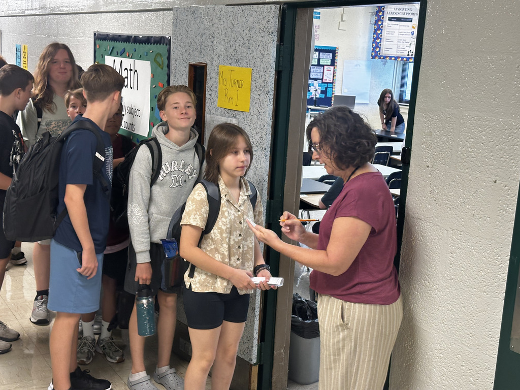 A teacher speaks to a girl in a school hallway; others, including a girl with a backpack, listen.