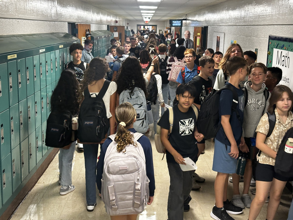 A hallway with lockers has a crowd of students, some carrying backpacks, lined up on either side.