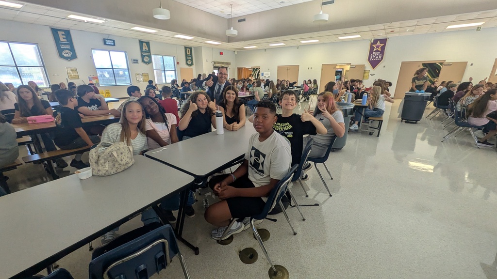 Classroom filled with students seated at desks, one student standing, with tables and chairs. White walls and floor.