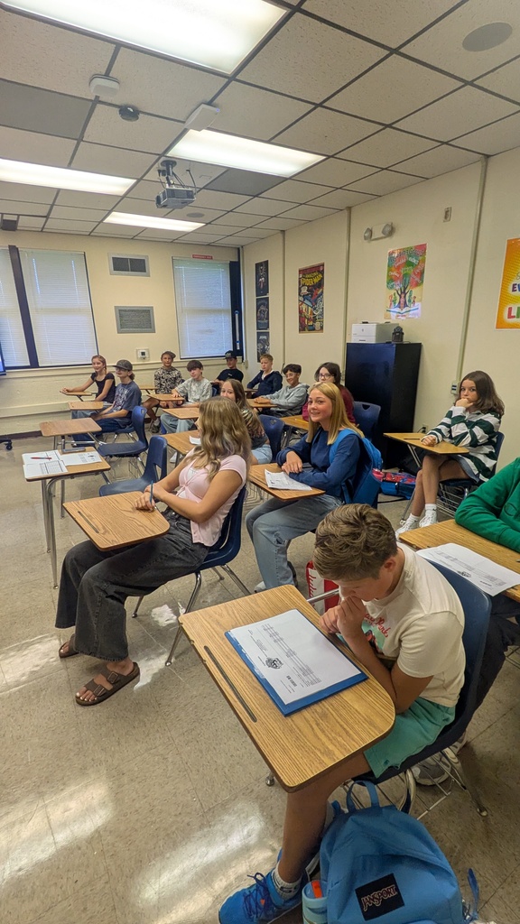 Students sit in desks, facing forward in a classroom. The room has white walls, windows, and overhead lights.