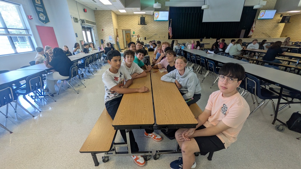 Group of students sitting at a long table in a cafeteria with many others sitting at tables.