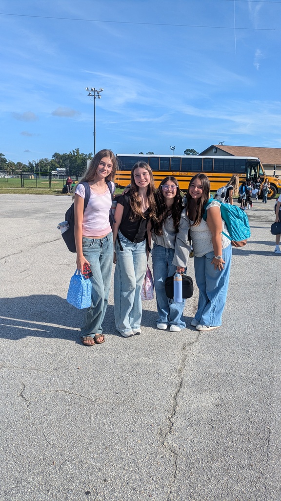 Four women in jeans stand in a parking lot with a yellow school bus in background.