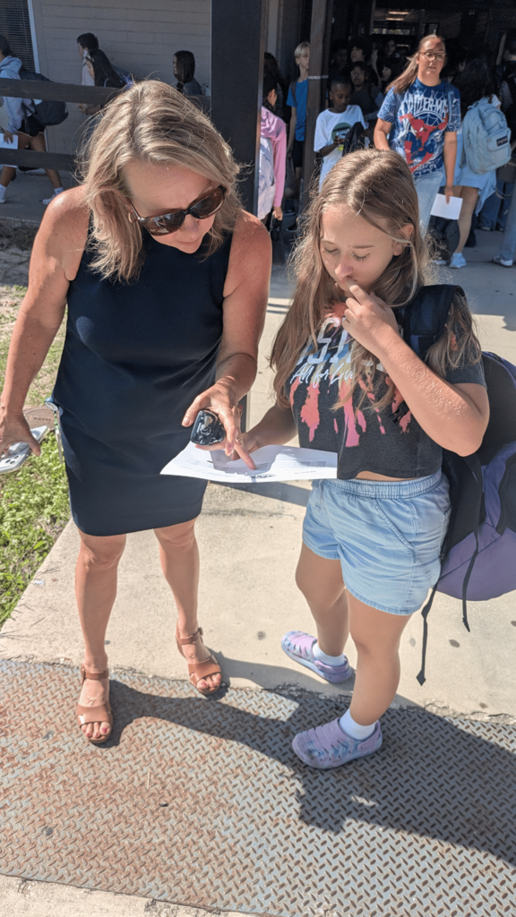 Two females, one with sunglasses and one with a backpack, examine a paper outdoors. Behind them, a building with a fence.