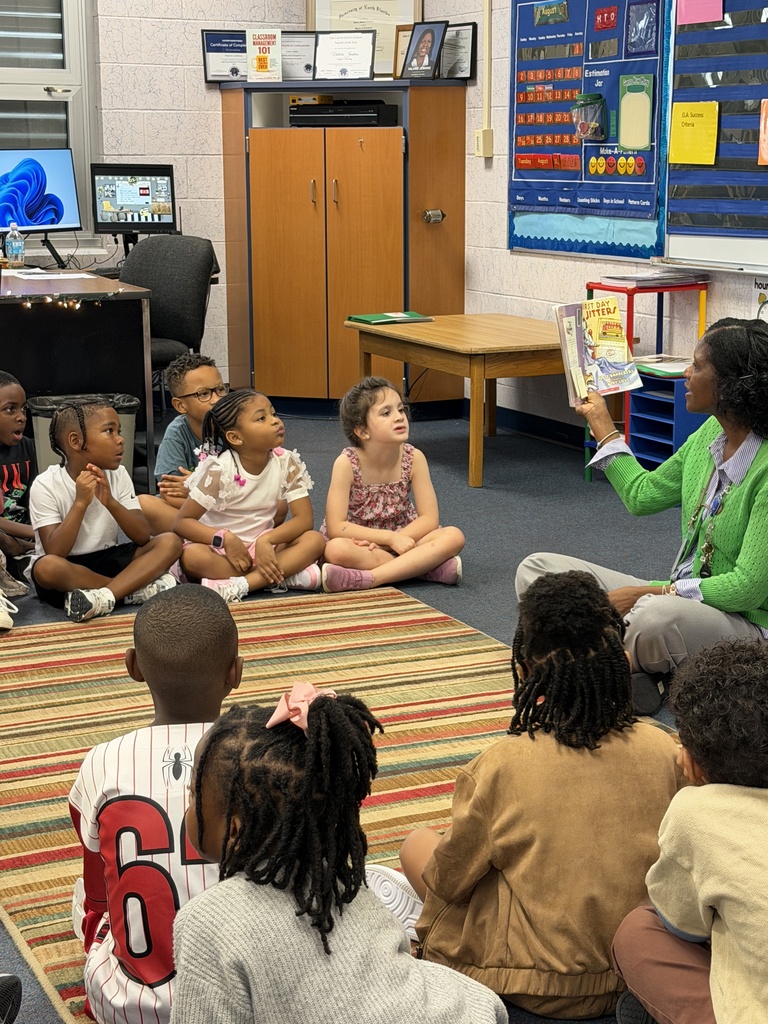 A group of children listen to a woman reading a book in a classroom. The room has a wooden table and rug.
