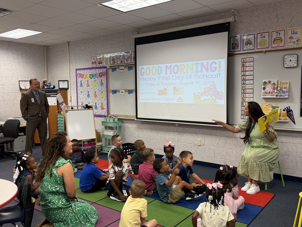 In a classroom, a woman reads to children sitting on the floor. A projector screen displays "GOOD MORNING!"