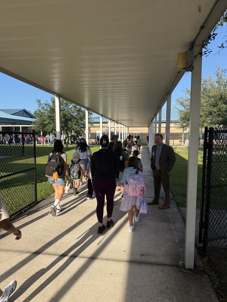 People walking on a covered walkway, some wearing backpacks. A man in a suit stands near a gate.