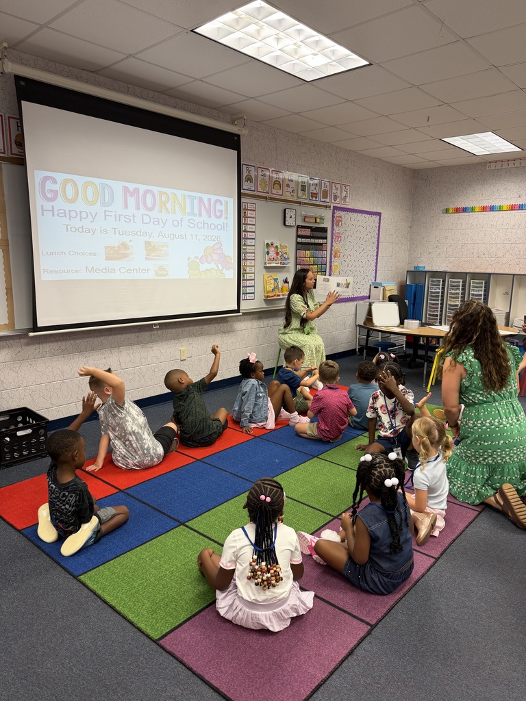 A classroom with children seated on the floor. A teacher stands in front of a projector screen showing the words "Good Morning."