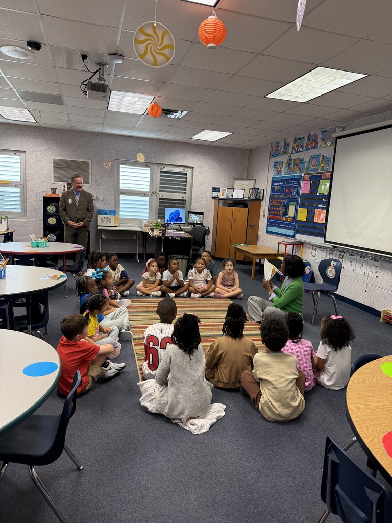 Classroom with children seated in a circle, a teacher reading aloud. Brightly lit room with tables, chairs, and a projector screen.