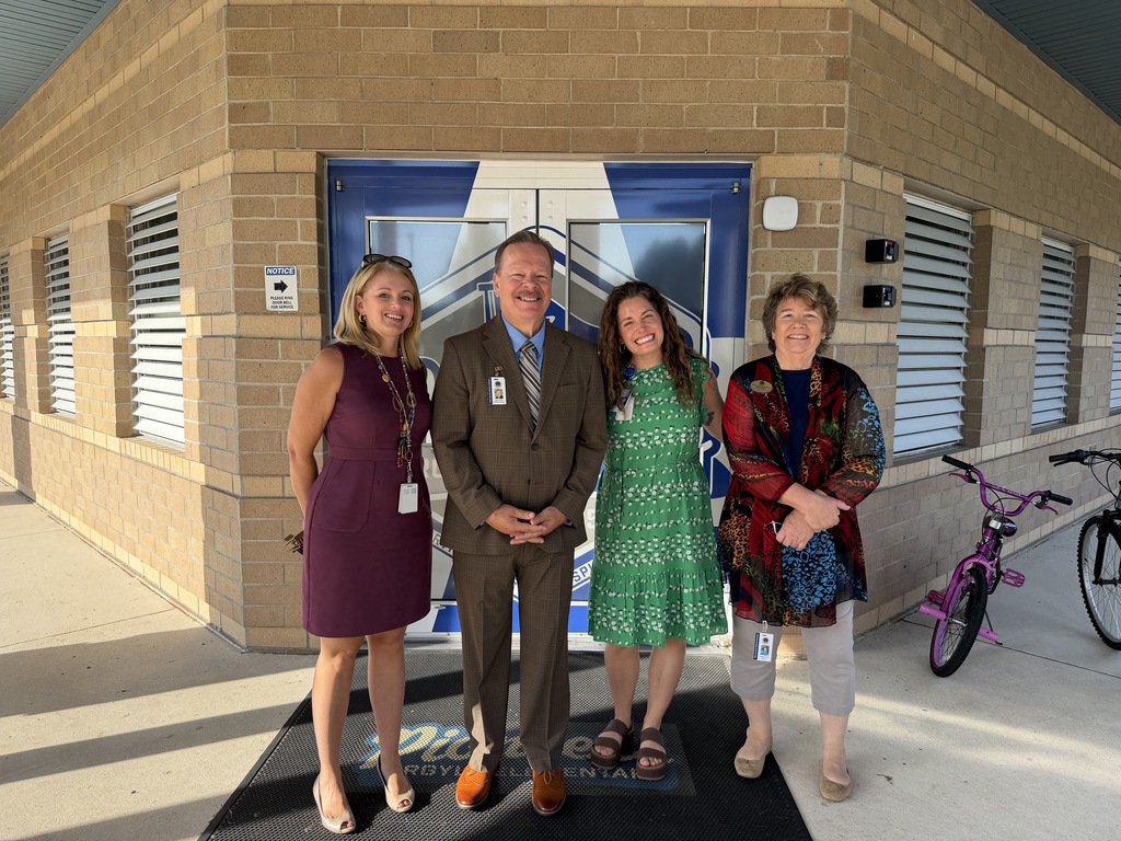Four people stand in front of a building with a blue door, smiling. A purple bicycle rests nearby.