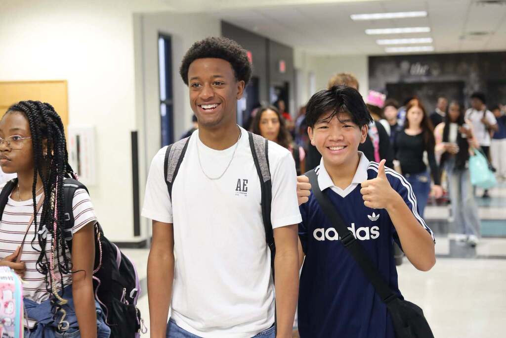 Two students standing in a school hallway, one smiling, the other giving a thumbs up. Background shows a busy corridor.