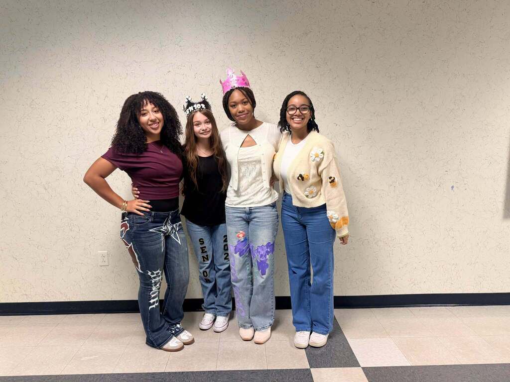 Four women pose together in a room. They are all standing with a white wall behind them.