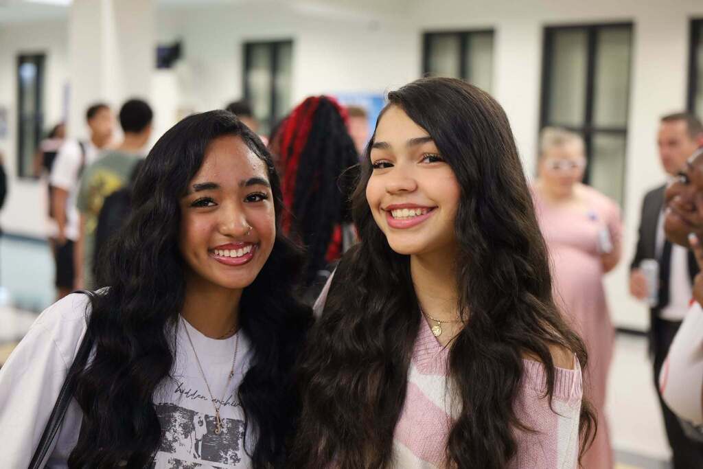 Two women with long hair, smiling, and posing for a photo in a hallway with other people.
