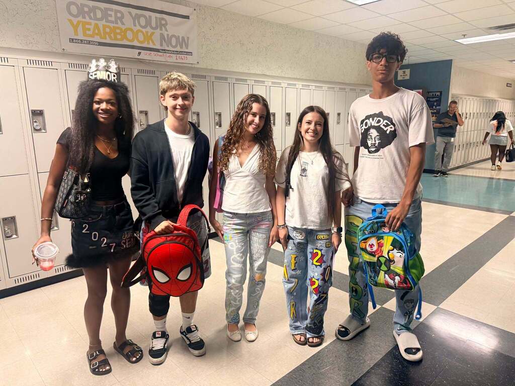 Group of five young adults standing in a school hallway, smiling, with lockers in the background.