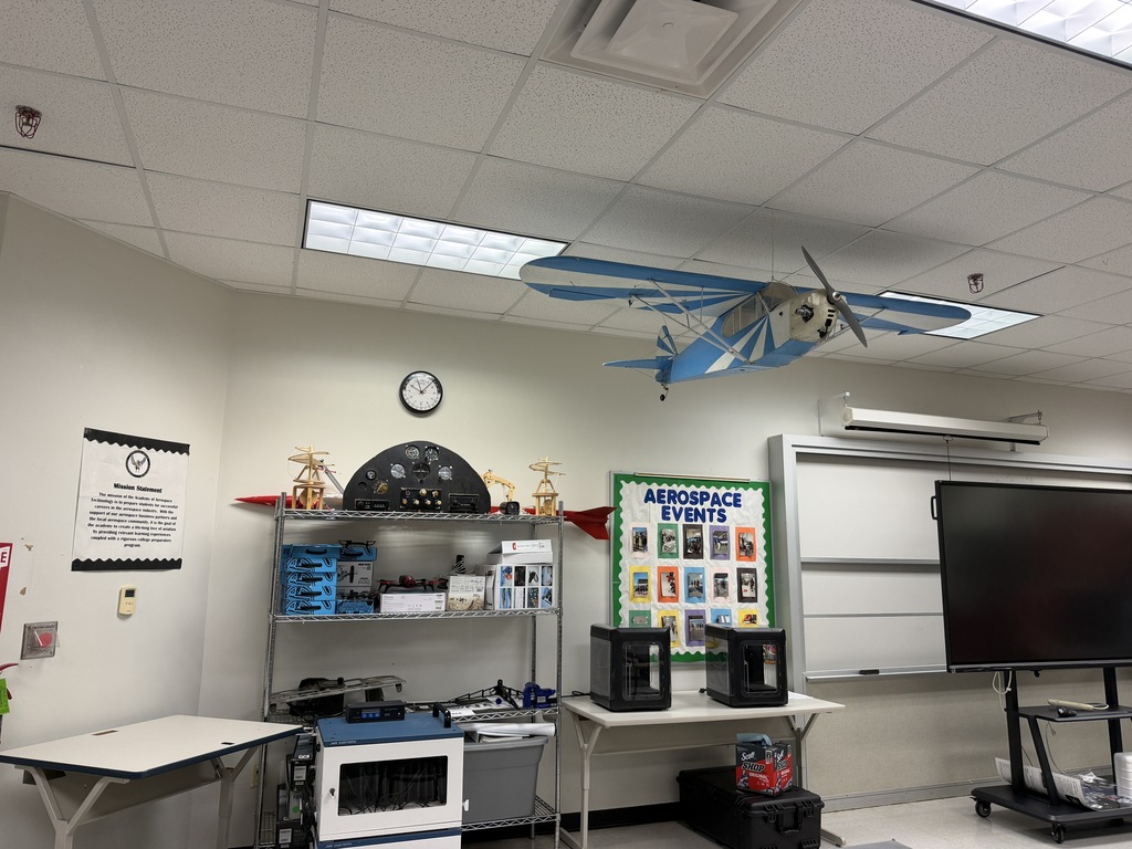 A classroom with a white ceiling, walls, and tables. An airplane is mounted on the ceiling. A whiteboard is mounted on the wall.